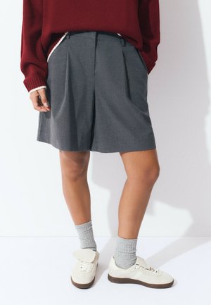 Persona vestida con pantalones cortos grises con pliegues, calcetines grises, zapatillas blancas con detalle de solapa y un suéter rojo, de pie contra un fondo blanco.