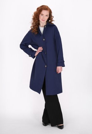 Marineblauer langer Mantel mit Kragen, sieben Knöpfen und hochgekrempelten Ärmeln. Getragen über schwarzen Hosen, die eine weiche Textur und eine taillierte Silhouette aufweisen.