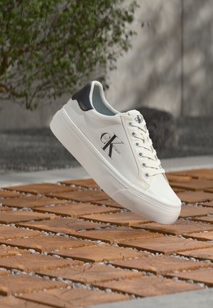 Biały sneaker Calvin Klein z czarną wkładką na pięcie i logo, pokazany w powietrzu nad drewnianą i kamienną nawierzchnią na zewnątrz.