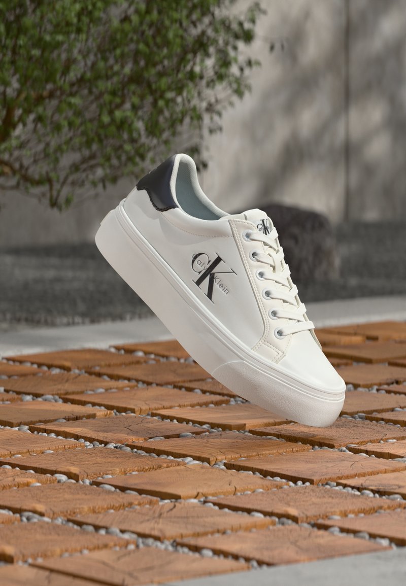 Weißer Calvin Klein Sneaker mit schwarzem Fersenlaschen und Logo, schwebt in der Luft über einem im Freien befindlichen Boden mit Holz- und Steindekor.