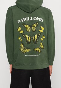 Zelená mikina s kapucí, na které je grafika motýla v žluté a černé barvě, s texty "PAPILLONS" a "Botanická výstava" níže. Bavlněná látka.