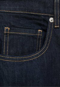 Jeans de mezclilla oscura con un bolsillo trasero que presenta costuras naranjas visibles y un remache de metal plateado. El material tiene un acabado suave y texturizado.