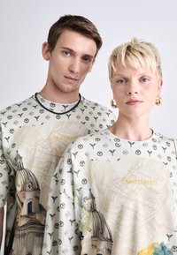 Overdimensionerede bomulds T-shirts med en lys cremefarvet bund, intrikate sorte mønstre og arkitektoniske tryk i bløde farver. Teksten lyder "CARLO COLUCCI."