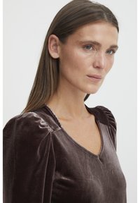Sametový top v tmavohnedej farbe s výstrihom do V, vyp puffovými rukávmi a hladkou textúrou s jemným leskom, ktorý zdôrazňuje elegantné konštrukčné detaily.