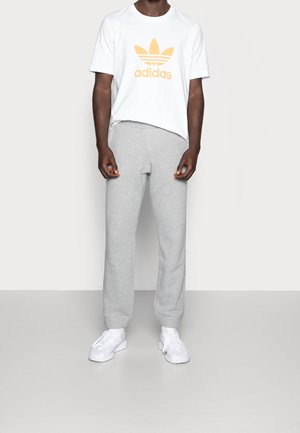 Osoba w białej koszulce Adidas z złotym logo, szarych spodniach dresowych i białych trampkach stoi na tle jednolitego tła.