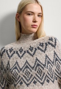 Maglione lavorato a maglia con colletto alto, base beige e motivi geometrici neri. La texture è morbida e spessa, sottolineando calore e comfort.
