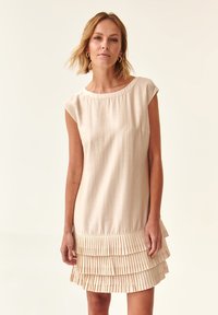 Knee-length vaaleancremivät dressi lyhyillä hihoilla, jossa on teksturoitu poimutettu röyhelö helman kohdalla. Sileä kangas pyöreällä pääntiellä.