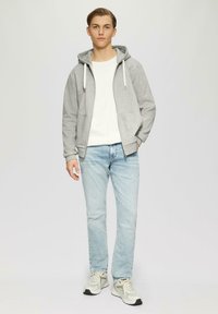 Grijze zip-up hoodie met trekkoorden, witte crewneck t-shirt, lichtblauwe spijkerbroek en grijze sneakers met zwarte accenten. Casual fit.