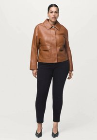 Chaqueta corta de cuero marrón con un gran cuello, dos bolsillos frontales de parche y cierre a presión. Combinada con jeans ajustados negros y tacones de punta.