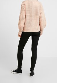 Person i en tjock ljusrosa stickad tröja, svarta skinny jeans och svarta high-top sneakers, stående vänd mot en enkel vit bakgrund.