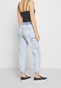 Kobieta w jasnoniebieskich spodniach cargo z wysokim stanem i elastycznymi mankietami, czarnej bluzce na cienkich ramiączkach i czarnych sneakersach wsuwanych, stojąca bokiem.