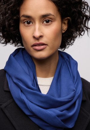 Écharpe bleue en tissu doux, drapée lâchement autour du cou, associée à un manteau noir et un haut à texture légère en dessous.