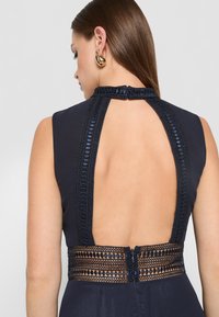 Zadný pohľad na bezrukávové námornícke šaty s čipkovaným odhaleným chrbtom a textúrovaným čipkovým pásom so zapínaním na zips.