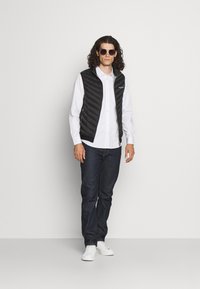 Svart quiltad väst över en vit skjorta med knapp, kombinerad med mörka denimjeans och vita sneakers; bärs med mörka solglasögon.