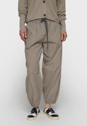 Person iført beige joggebukser med elastisk talje og sidelommer, en beige knappet cardigan og sort-hvide sneakers.