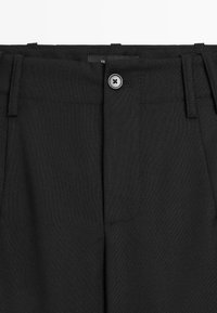 Gros plan sur la ceinture d’un pantalon noir avec passants de ceinture et une fermeture par un seul bouton au centre devant.