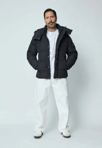 Doudoune noire à capuche, fermeture par boutons et motif matelassé, portée sur un t-shirt blanc et associée à un pantalon blanc déchiré.