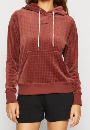 Vrouw draagt een roestkleurige, gestructureerde Nike-hoodie met witte trekkoorden en zwarte korte broek, afgebeeld van nek tot halverwege het dijbeen.
