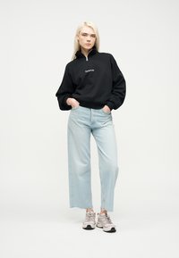 Ξανθιά γυναίκα που φορά μαύρο φούτερ Napapijri με φερμουάρ στο μισό μήκος, ανοιχτό μπλε τζιν φαρδύ στις μπάλες και μπεζ αθλητικά παπούτσια, όρθια με τα χέρια στις τσέπες.