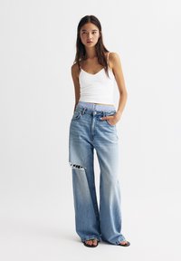 Light blue wide-leg jeans with a distressed tear, paired with a white fitted tank top. The jeans feature a contrasting waistband detail.