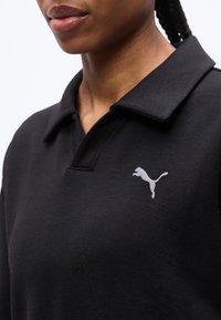 Κοντινό πλάνο ατόμου που φοράει μαύρο πουκάμισο με γιακά και λευκό λογότυπο Puma στο αριστερό στήθος, με πλεξούδες που φαίνονται από πίσω.