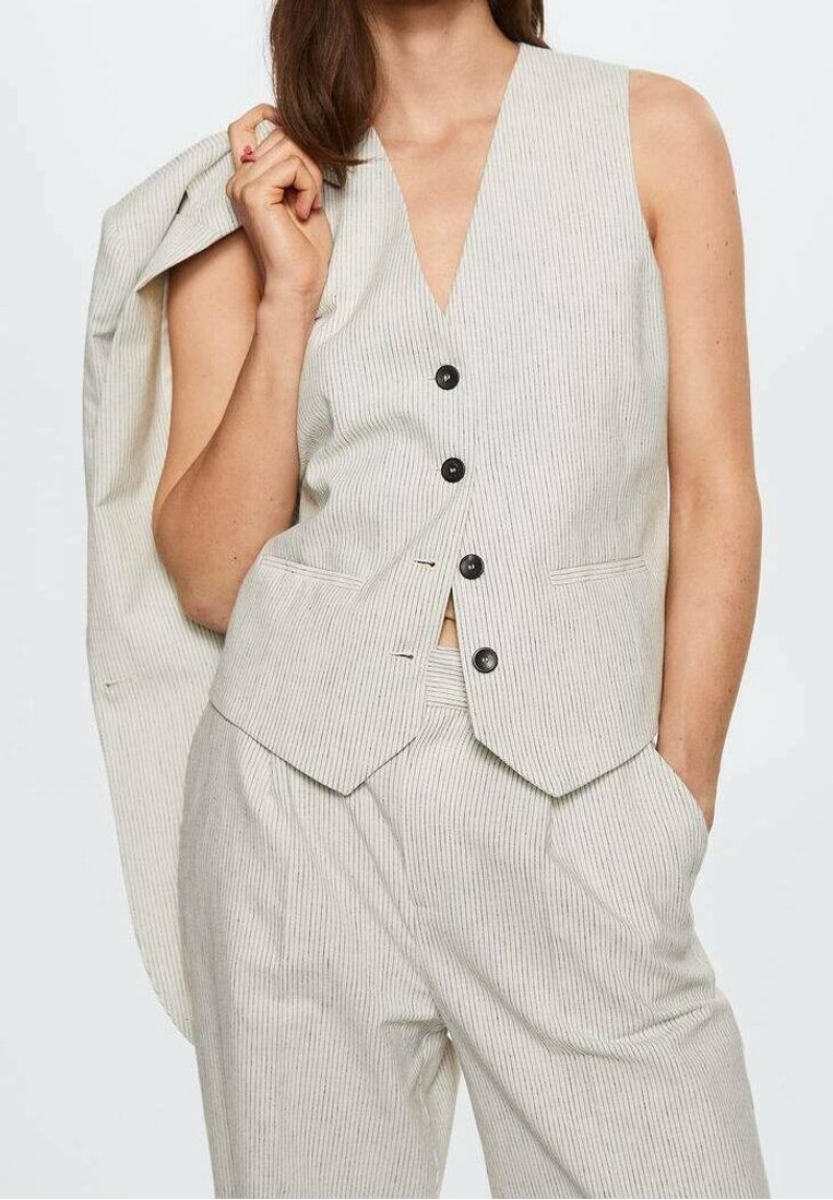 Femme portant un gilet sans manches beige à fines rayures et un pantalon assorti, tenant une veste sur une épaule avec une main dans la poche.