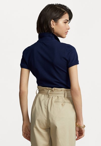 Jūrininkų polo marškiniai tamsiai mėlynos spalvos, su aukštu apykakle, trumpomis rankovėmis ir prigludusiu siluetu, derinami su aukšto juosmens kreminės spalvos kelnėmis, turinčiomis mygtukų akcentą.