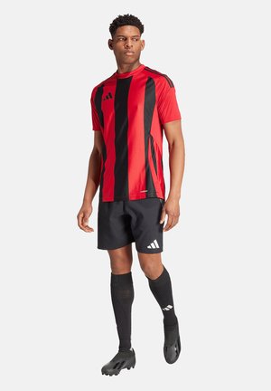 Mann trägt rot-schwarz gestreiftes Fußballtrikot, schwarze Shorts, schwarze knielange Socken und schwarze Fußballschuhe, steht und schaut zur Seite.