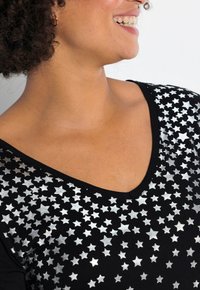 Schwarzes Top mit V-Ausschnitt, mit einem Muster aus glänzenden silbernen Sternen, die sich über den Stoff verteilen und eine glatte Textur hervorheben.