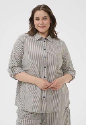 Lachende vrouw met golvend bruin haar, gekleed in een grijs-wit verticale gestreepte blouse met knopen en bijpassende broek, handen voor zich gevouwen.