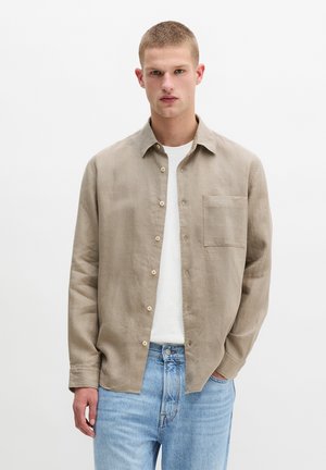 Junger Mann trägt beige Leinen-Hemd über weißem T-Shirt und hellblauen Jeans, steht vor einem schlichten weißen Hintergrund.