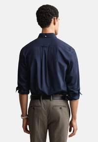 Camisa azul-marinho com botões, mangas arregaçadas, tecido texturizado e um pequeno botão na gola, combinada com calças cinzentas estampadas.