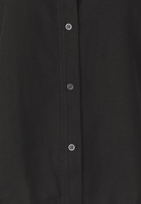 Chemise noire à boutons avec un tissu texturé, comportant six boutons sur le devant, avec des boutons en plastique gris et un col classique.