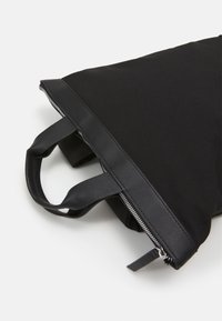 Sac fourre-tout en tissu noir avec poignées en cuir, présentant une surface lisse, une fermeture éclair en haut et des accents de design minimalistes.