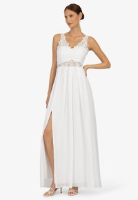 Witte jurk met een kanten lijfje, bloemenpatronen en een hoge split. De rok is zwierig, aangevuld met strappy witte hakken.