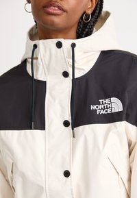 Bijela i crna vodootporna jakna s kapuljačom, koja ima čvrsti crni gornji dio, kopčanje na dugmad i logotip The North Face. Teksturirani materijal.