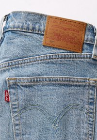 Pantalones vaqueros Levi's 501 de mezclilla azul con el bolsillo trasero que tiene una etiqueta roja de Levi's y un parche de cuero marrón de la marca cosido encima del bolsillo.