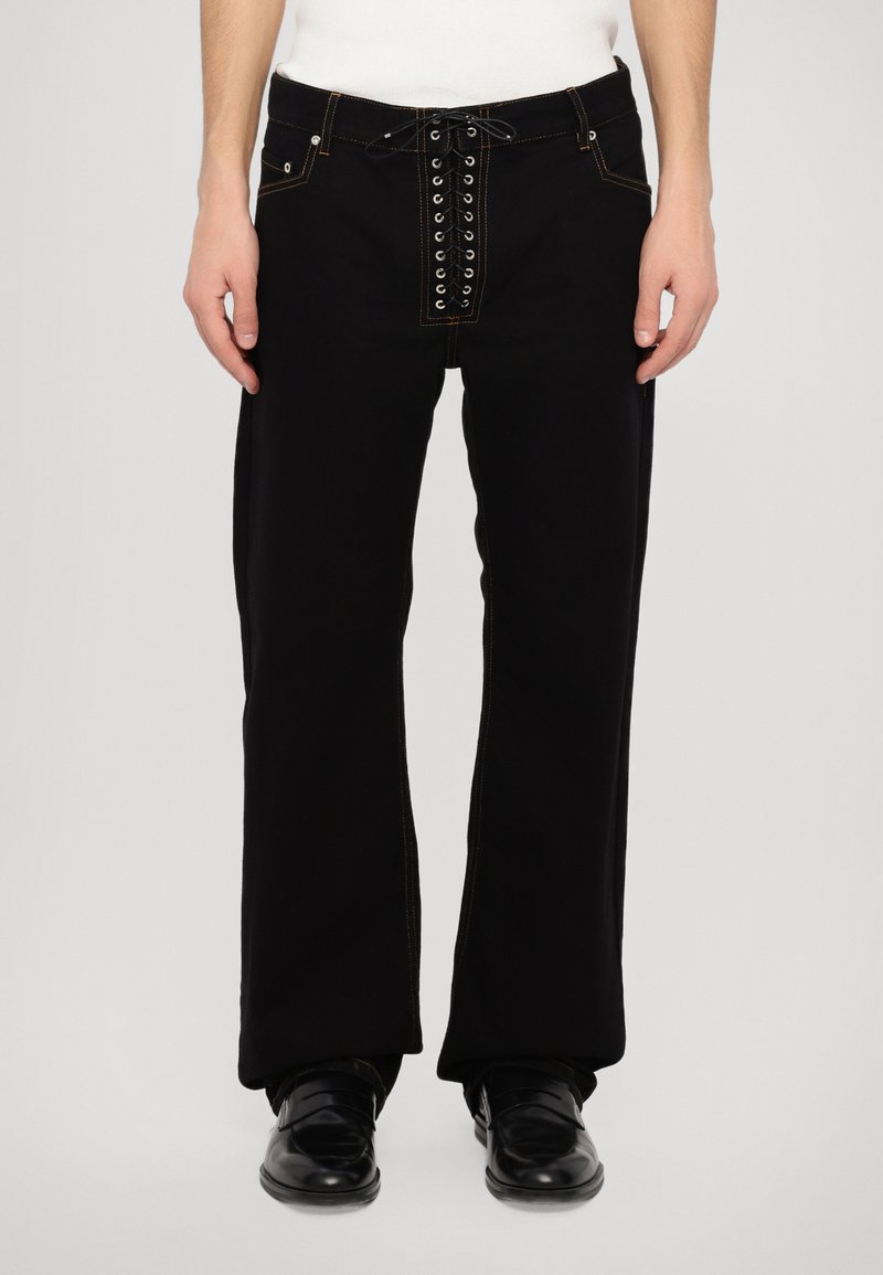 Black pants with a loose fit, featuring a front lace-up design with metal grommets and contrasting orange stitching. Flat pockets visible.