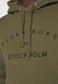 Sweat à capuche vert olive avec du texte noir imprimé "BJÖRN BORG 1955 STOCKHOLM", comprenant une capuche avec cordon et un tissu texturé. Oeillets en métal pour les cordons de la capuche.