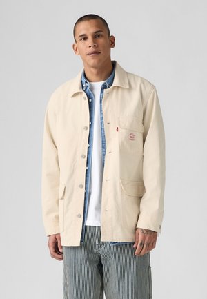 Beige katoenen werkjack met knoopsluiting, twee grote zakken en een klein logo op de borst. Draagt over een spijkerhemd en een wit T-shirt. Gestreepte broek.