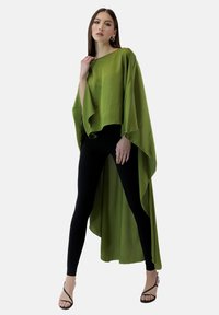 Zielona, zwiewna bluzka o asymetrycznym dolnym wykończeniu, wykonana z lekkiego materiału, w połączeniu z czarnymi legginsami i czarnymi sandałami na ramiączkach.