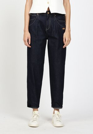 Person wearing dark blue high-waist relaxed-fit jeans with rolled cuffs, white sneakers, and a cropped white sleeveless top.