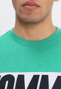 La camiseta de algodón verde presenta un escote redondo y un gran texto impreso en azul oscuro sobre un panel blanco horizontal en el pecho.