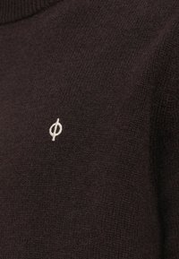 Sötétbarna kötött pulóver magas nyakkal és egy kis fehér logóval a mellkason, texturált anyagból.