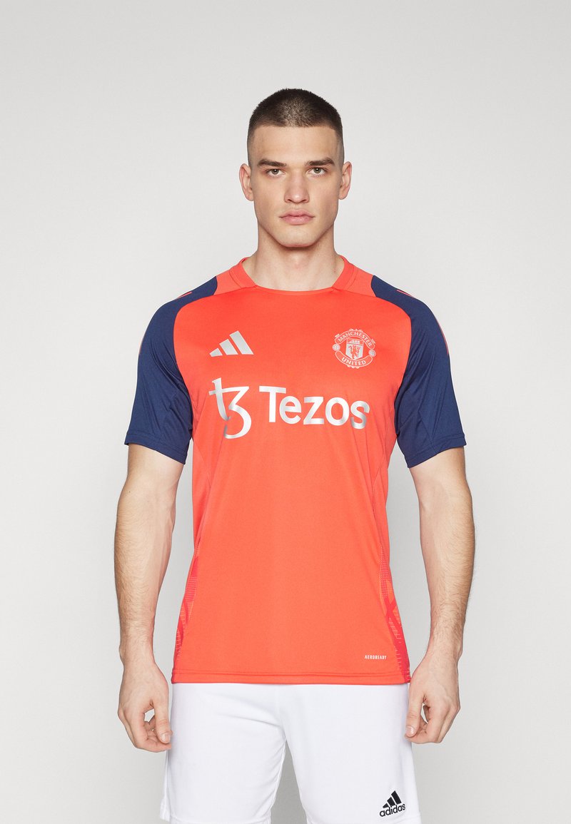 Sarkans un tumši zils sporta krekls ar īsām piedurknēm, ar Manchester United logo un "Tezos" uzdrukātu uz priekšpuses. Gluda auduma, ikdienišķs piegriezums.