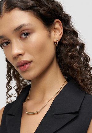 Gold necklace with curved bar pendant, worn over a black collared top. Curly hair, subtle earrings, and neutral lip color visible.
