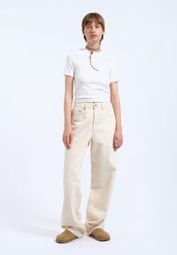 Witte, getailleerde t-shirt gecombineerd met lichtbeige wijde broek. De outfit heeft subtiele stikseldetails en een neutrale tint. Zachte, gestructureerde stof.