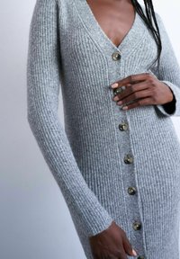 Personne à la peau foncée portant un long cardigan gris en maille côtelée, boutonné, à manches longues, touchant un bouton avec des ongles soignés.