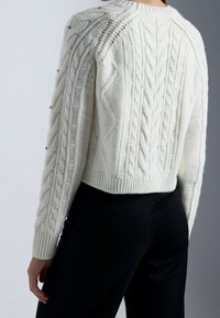 Weißer Kabelstrickpullover mit strukturierten Mustern und Rundhalsausschnitt, verziert mit dekorativen Knöpfen an den Ärmeln und einem Rippenbund.
