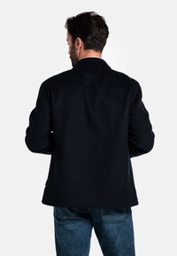 Manteau en laine noir avec un col classique, des manches longues et une coupe droite, porté sur un jean en denim bleu, vu de dos.